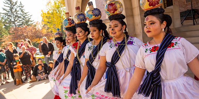 Image of a row of dancers with painted faces, balancing pottery on their heads. Vibrant colors and traditional costumes underline the celebration. Crowd of people watch from behind the dancers. Image from OPM&#39;s Dia de los Muertos Celebration in fall 2022.