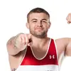 A person in a red-and-white wrestling singlet with "Wisconsin" and "B1G" logos poses confidently, flexing one arm and pointing forward. The background is plain white.