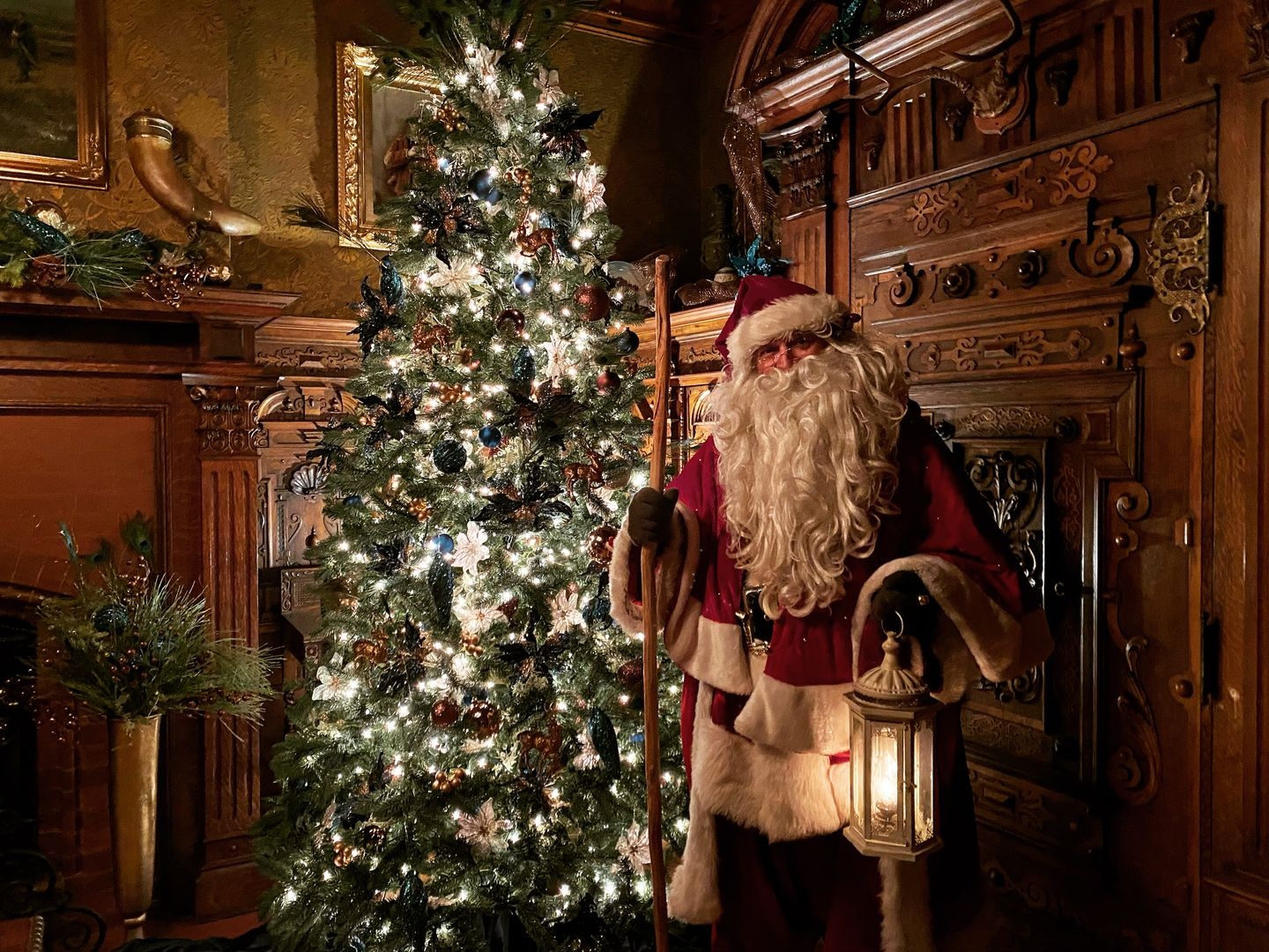 A person dressed as Santa Claus stands beside a beautifully decorated Christmas tree in an ornate, warmly lit room, holding a lantern and a staff.