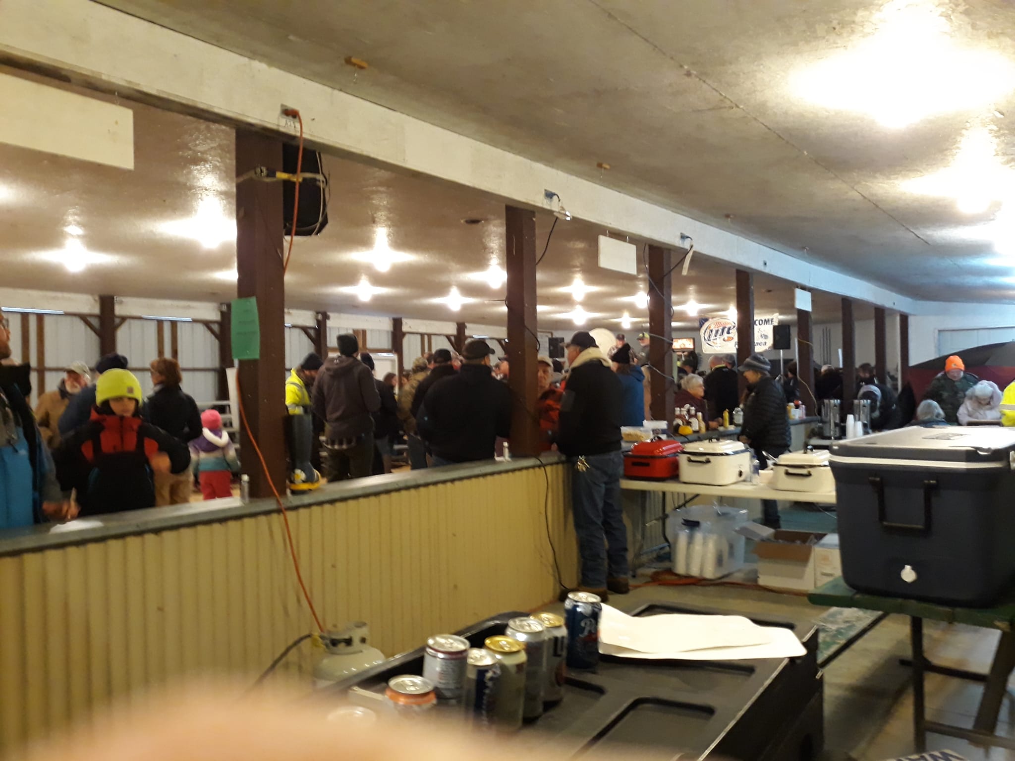 People of all ages gather inside a large, well-lit pavilion for a winter event. Attendees are dressed in warm clothing, and food stations with slow cookers, drinks, and supplies line the foreground.