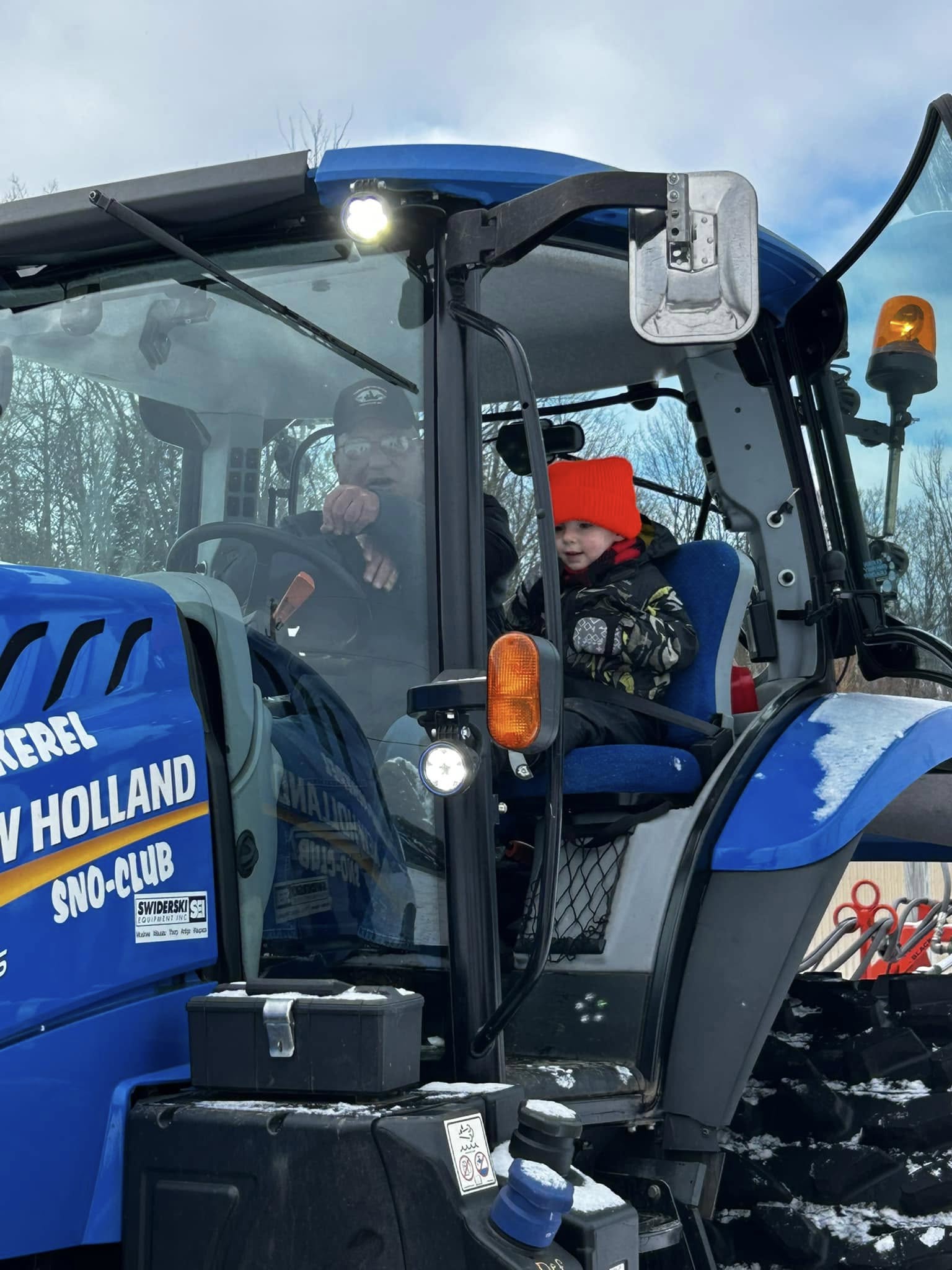 A child beams with excitement while sitting beside an adult in the cab of a Tombstone-Pickerel Sno-Club New Holland tractor. Bundled up in winter gear, the pair enjoys a close-up look at the snow grooming equipment during a fun, family-friendly winter event in Langlade County.