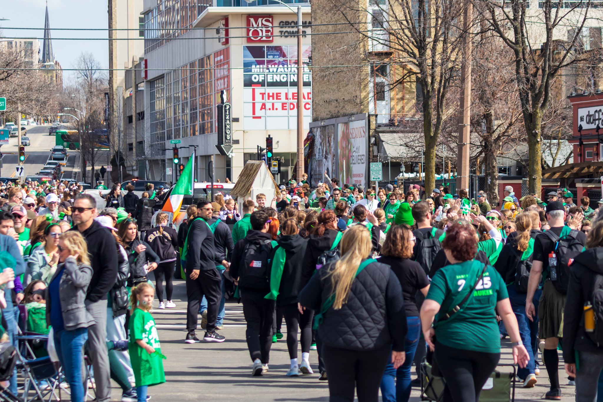 Crowds of people dressed in green gather along a city street during a festive parade, waving flags and enjoying the celebration.