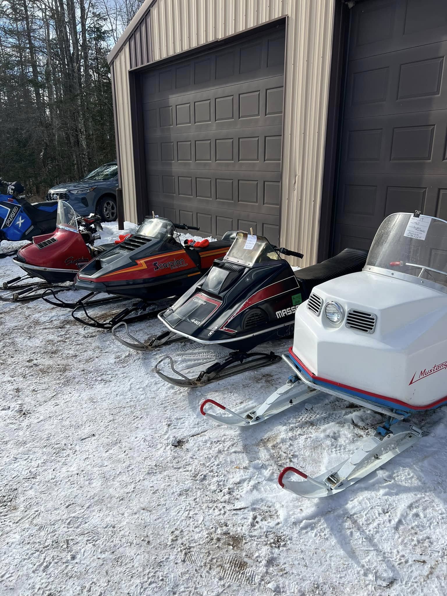 A row of well-preserved vintage snowmobiles, including classic models like Scorpion, Massey, and Mustang, is lined up outside a garage at the Tombstone-Pickerel Snowmobile Club event. Resting on a light blanket of snow, these nostalgic machines showcase the rich history and passion of snowmobiling in Langlade County.