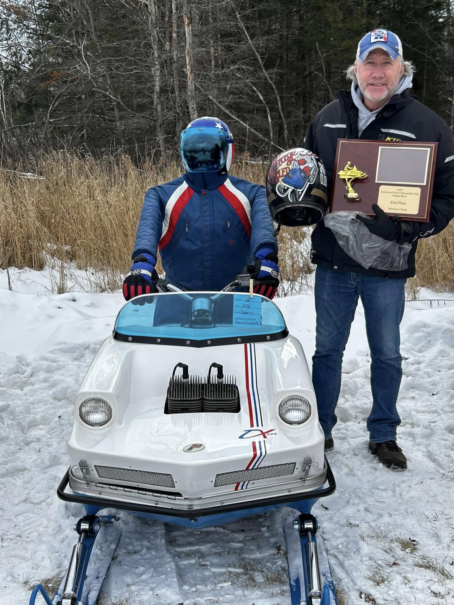 One person sits on a classic snowmobile in full retro riding gear, while another holds a helmet and a first-place plaque. Snow blankets the ground, and a backdrop of tall grass and forest adds to the nostalgic Northwoods winter setting.