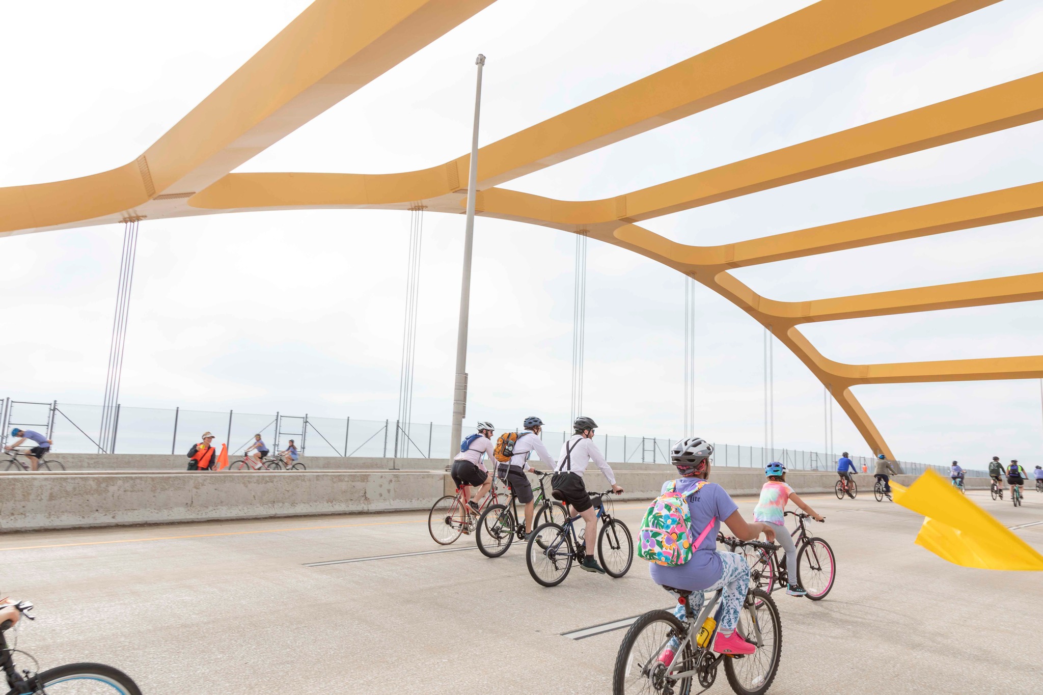 Cyclists ride along a lakeside path under large yellow shade canopies on a bright, sunny day.