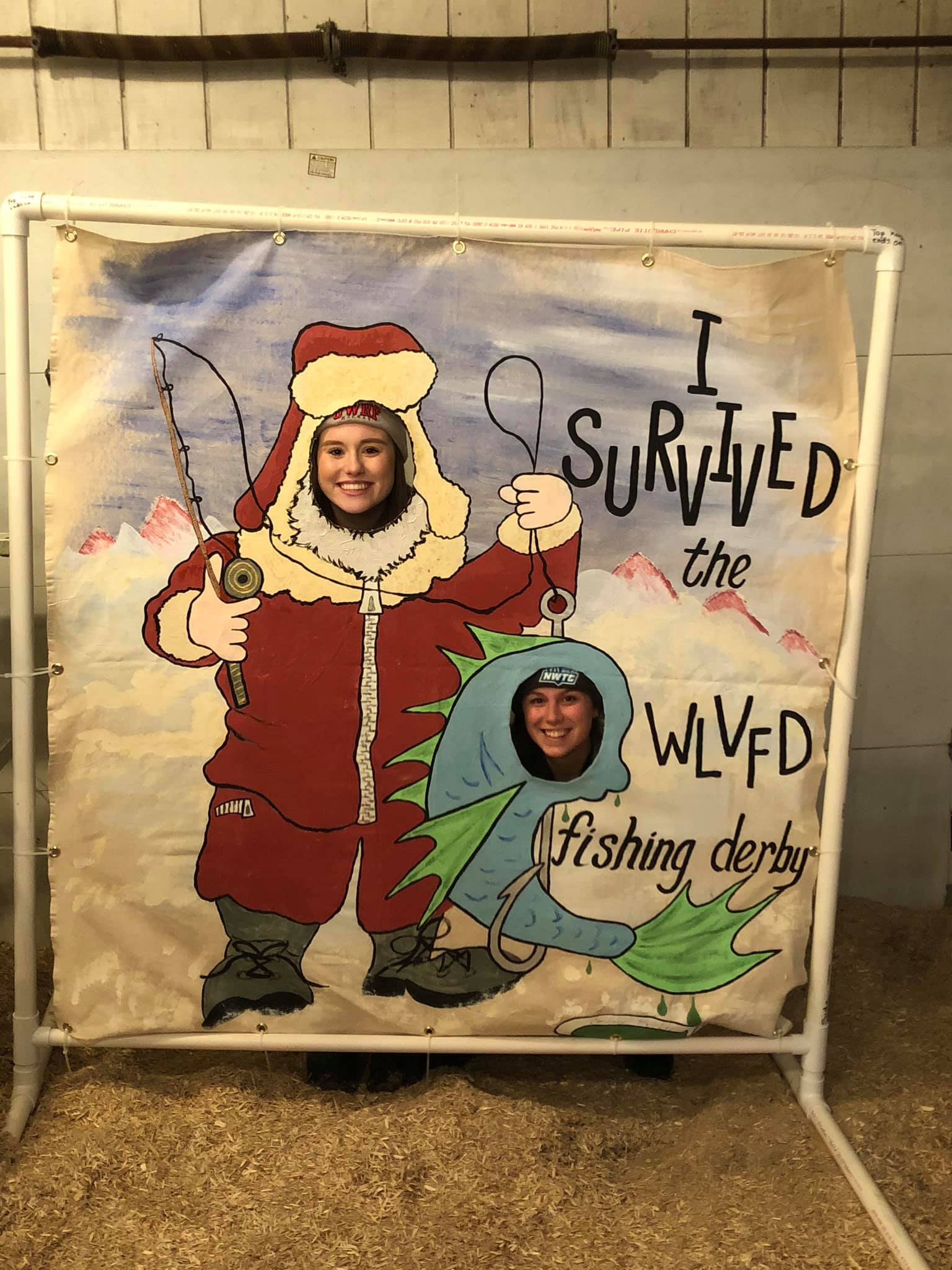 Two smiling participants pose behind a playful cutout sign that reads "I Survived the WLVFD Fishing Derby." One person appears as a bundled-up ice fisher and the other as a cartoon fish.