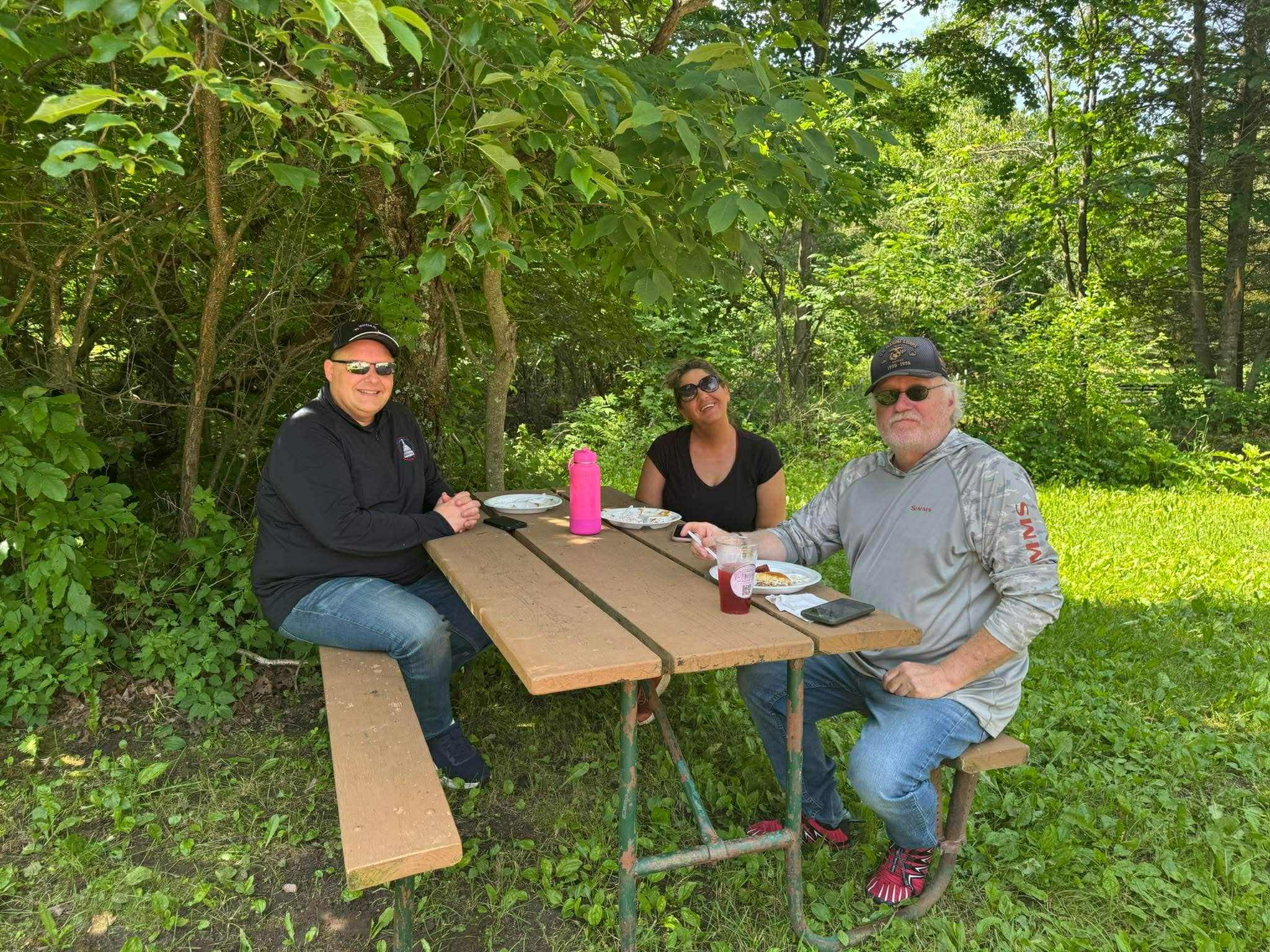 Three people enjoy a meal at a wooden picnic table nestled in a shaded grassy area surrounded by thick forest greenery. Plates and drinks are set out as they smile and relax in the peaceful, natural setting.