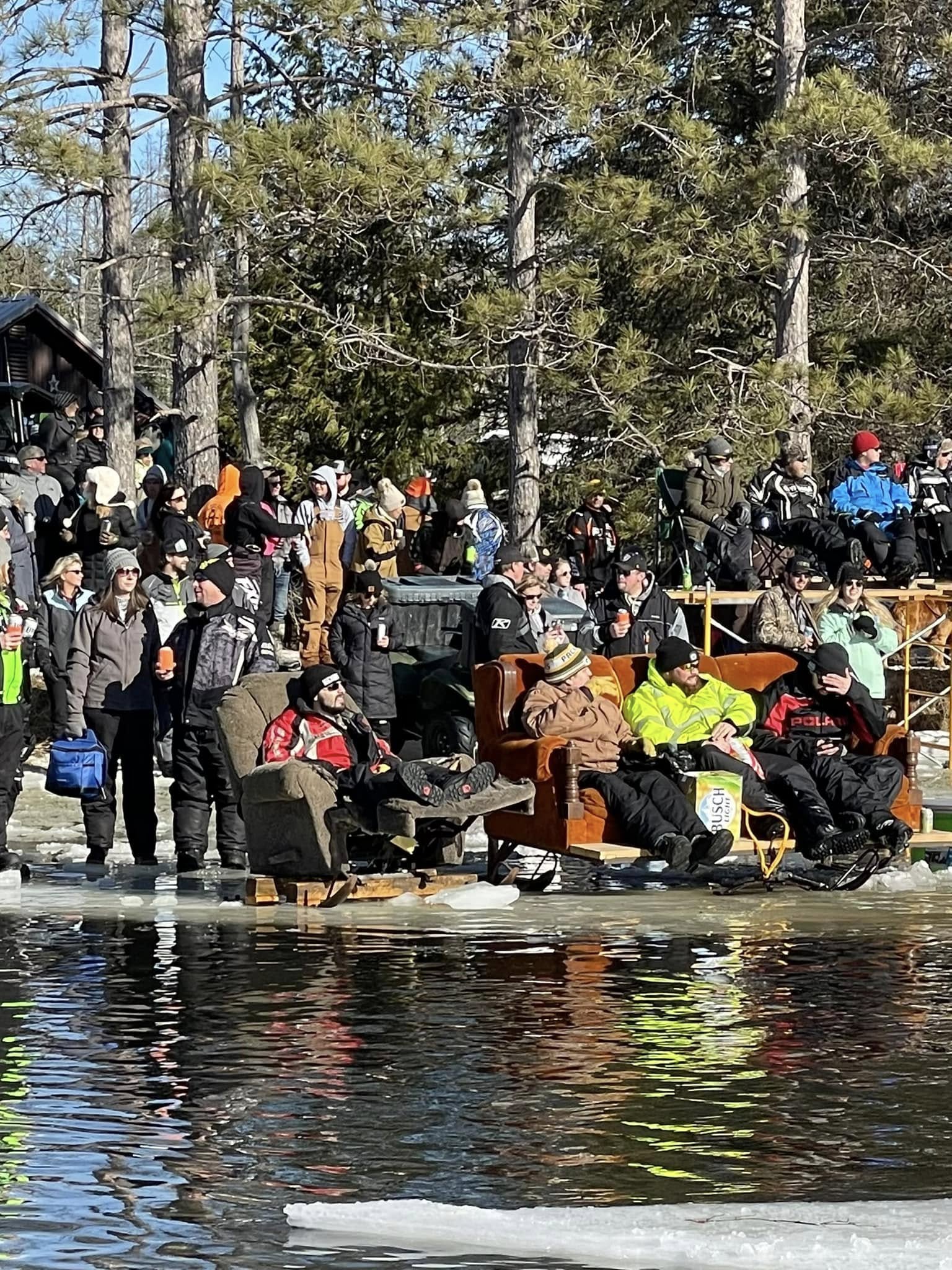 Dozens of people gather on the frozen shoreline, bundled in winter gear and enjoying a sunny winter day. A group relaxes in recliners and couches set directly on the ice, creating a playful and laid-back atmosphere as they overlook the partially open water. Tall pine trees and a rustic setting enhance the unique outdoor gathering.