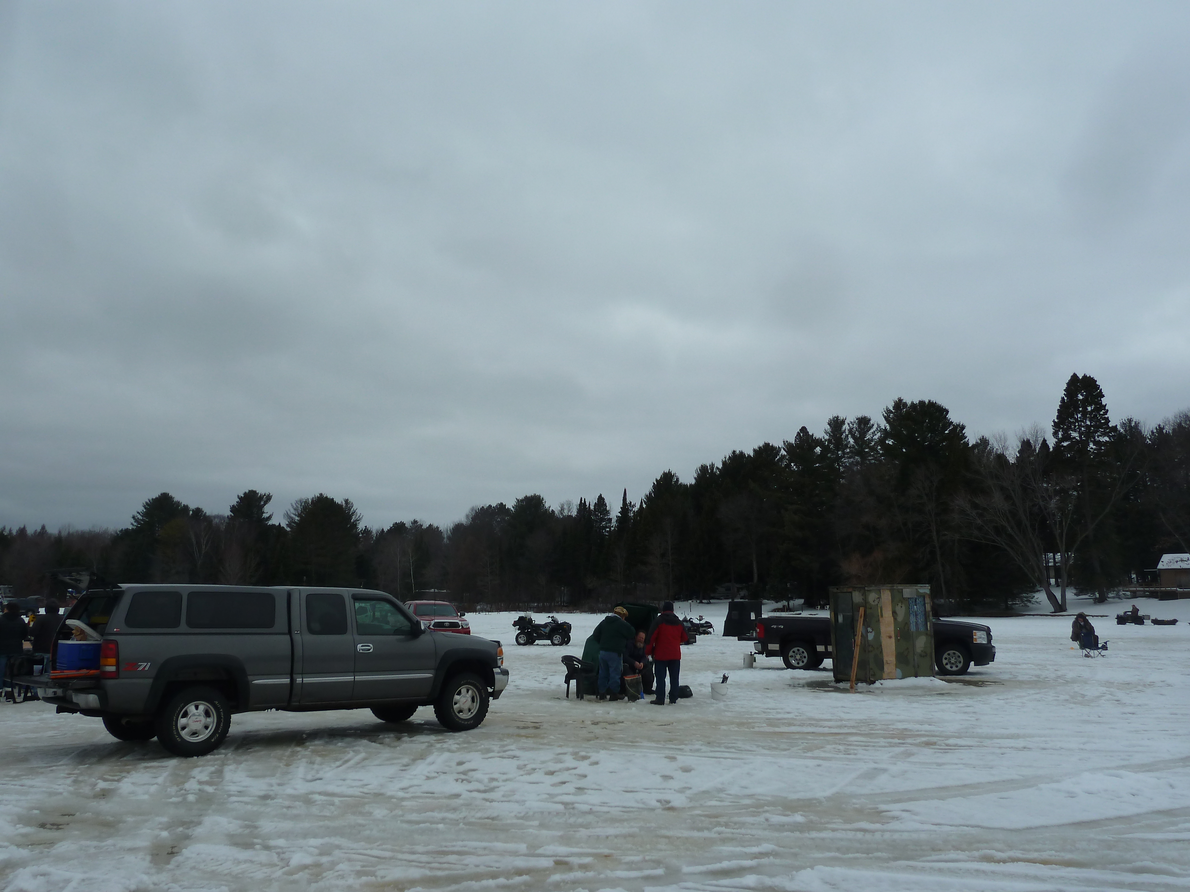 People gather near trucks and ice shanties on a frozen lake, preparing for a day of ice fishing under overcast skies. Snowmobiles and ATVs are scattered in the distance, while the tree-lined shore provides a scenic winter backdrop.