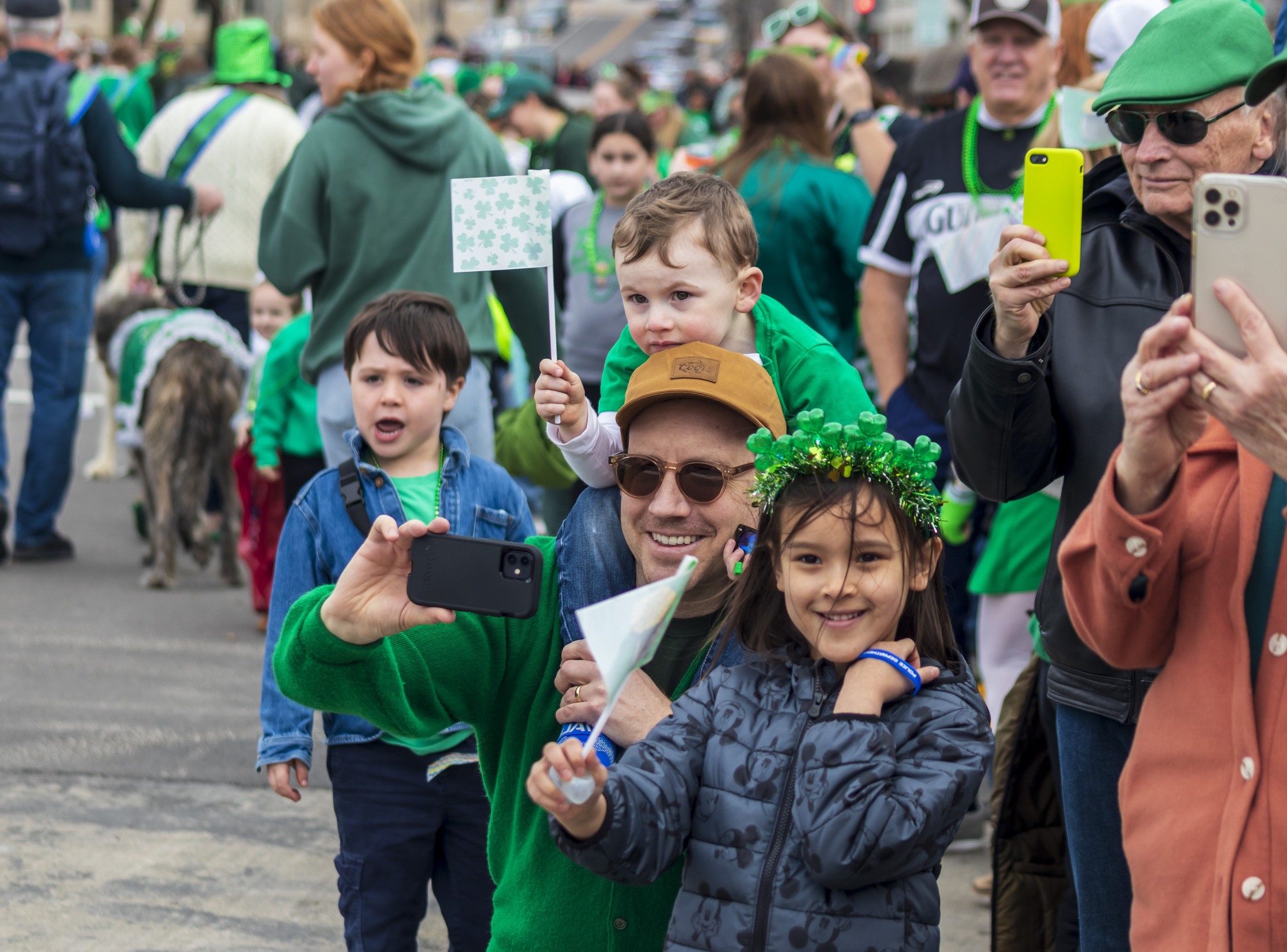 Smiling children dressed in green stand at the edge of a parade route, waving and holding festive accessories while people in green clothing walk by in the background.
