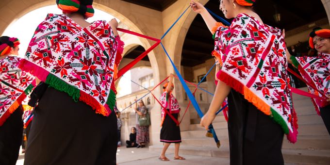 A group of dancers wearing bright costumes circle around each other, connected by fabric as part of their dance. They&#39;re wearing bright pink, orange, and green shawls over black skirts.