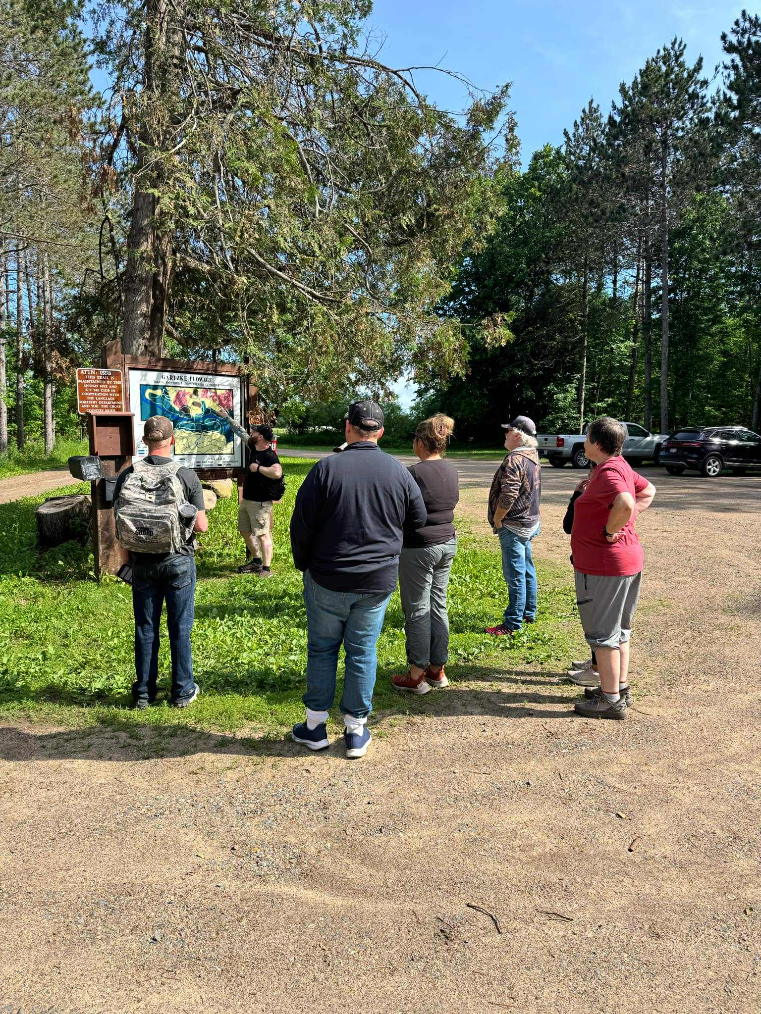 A small group of people gathers near a trailhead map and informational signs at Gartzke Flowage. Surrounded by tall pine trees, a guide points at the map, explaining the hiking route. Vehicles are parked nearby under a clear blue sky, signaling the start of an outdoor adventure.