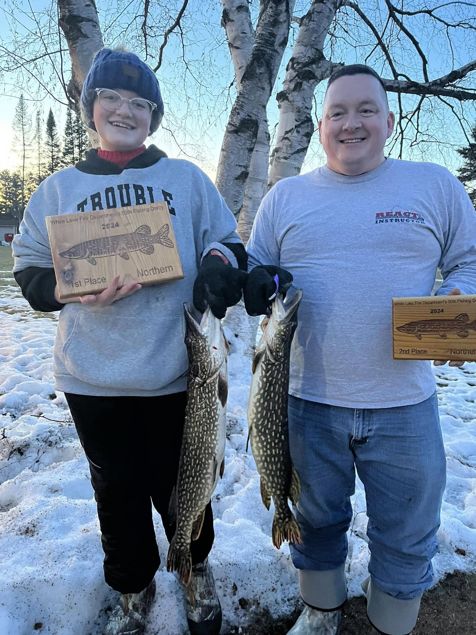Two smiling participants proudly display their catches and wooden award plaques after the winter fishing derby. Standing in snow with northern pike in hand, they celebrate 1st and 2nd place wins