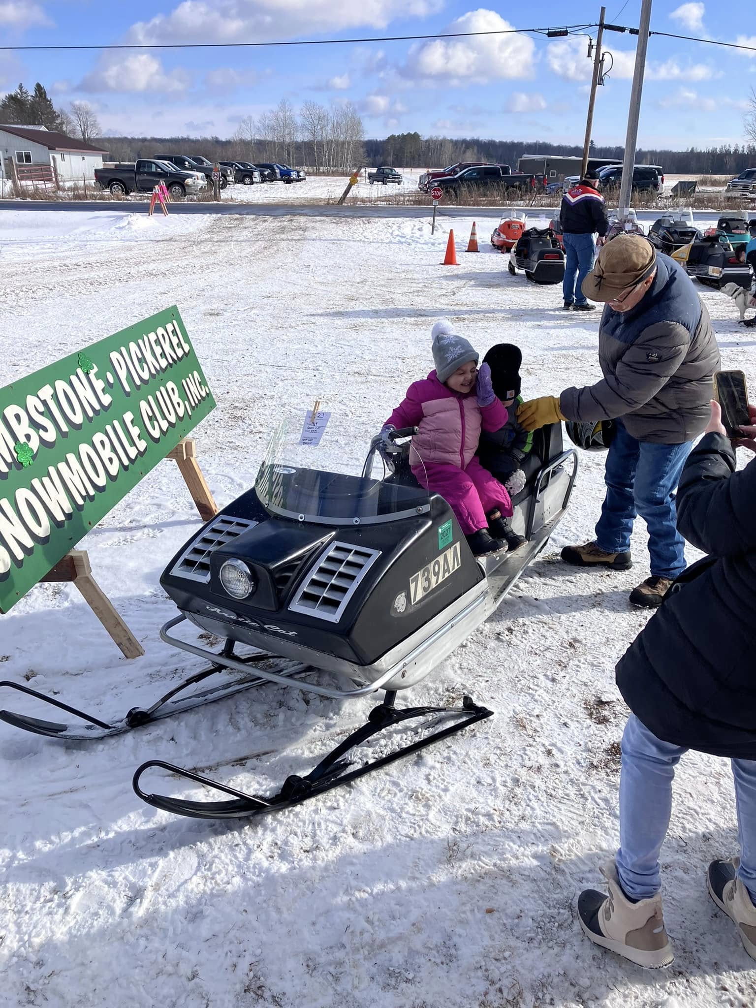 A joyful moment unfolds at the Tombstone-Pickerel Snowmobile Club event as a smiling young girl, bundled up in winter gear, sits on a vintage snowmobile. An older gentleman helps her get settled while family members capture the memory.