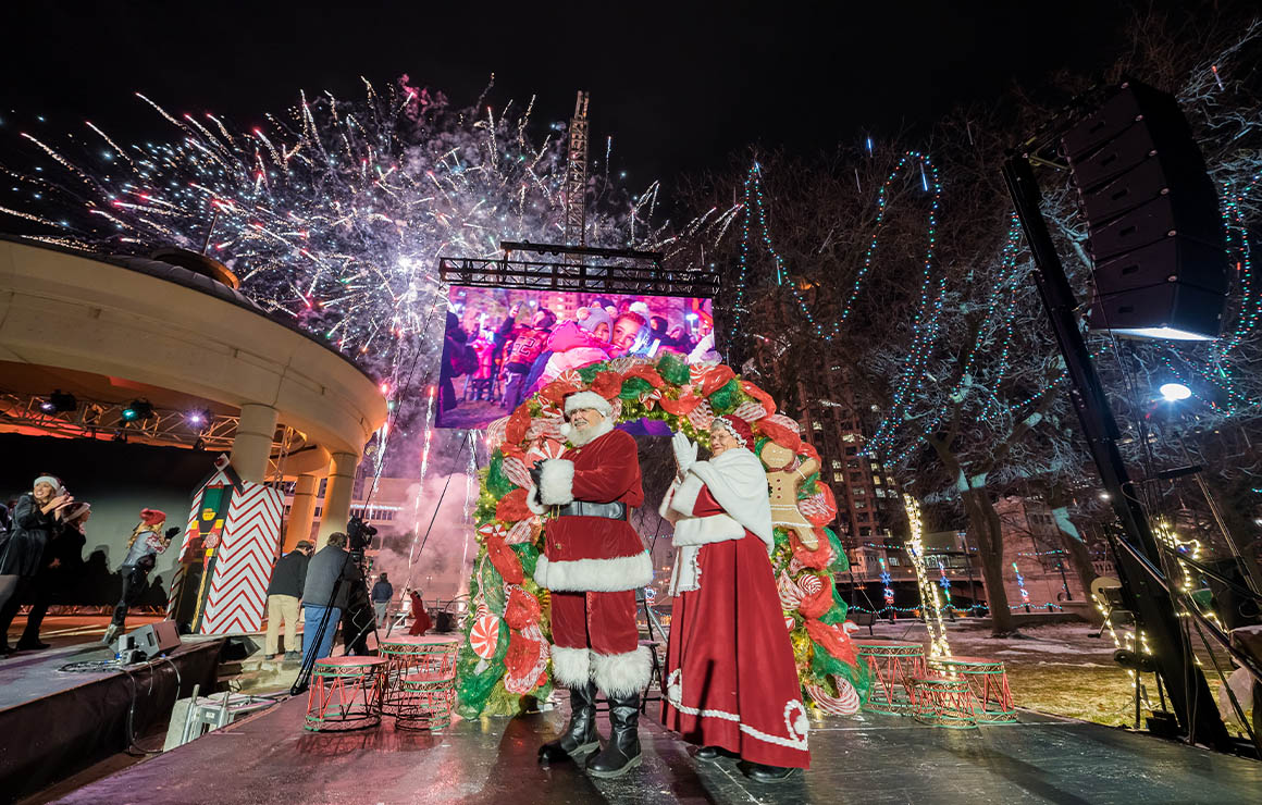 Santa and Mrs. Claus greet visitors in a festively decorated outdoor plaza as fireworks light up the night sky and crowds gather to celebrate.