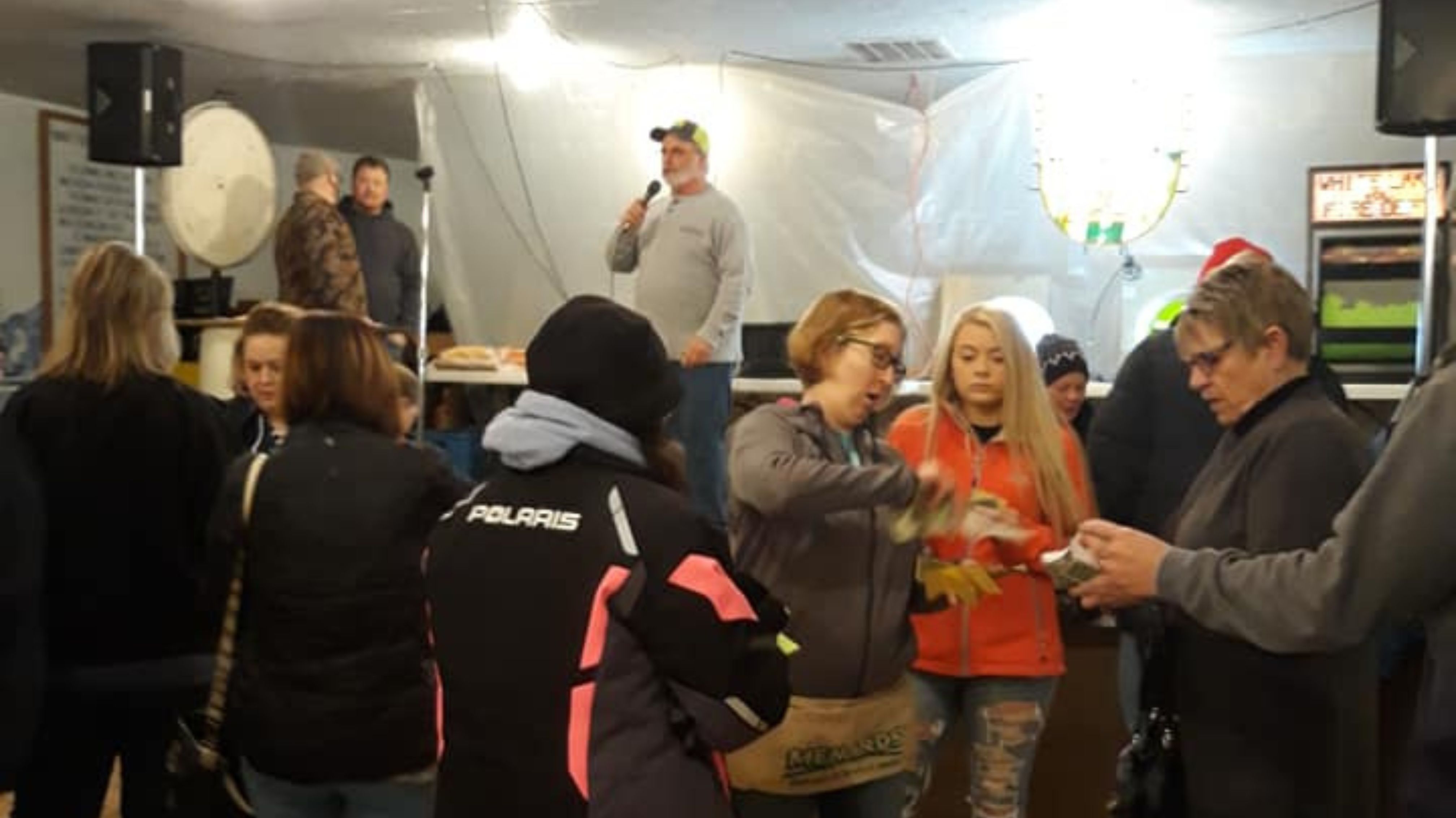 A crowd gathers indoors, where a man speaks into a microphone near a prize table. Attendees chat, count raffle tickets, and enjoy the lively atmosphere.