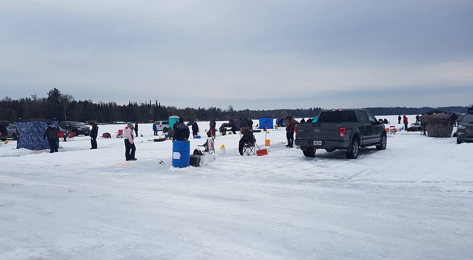 Participants gather on a frozen lake for a winter fishing event, with trucks, ice shanties, and portable shelters spread across the snow-covered surface. People are bundled in cold-weather gear, tending to fishing holes and enjoying the outdoor experience beneath a cloudy sky. The surrounding forest adds a natural backdrop to the scene.