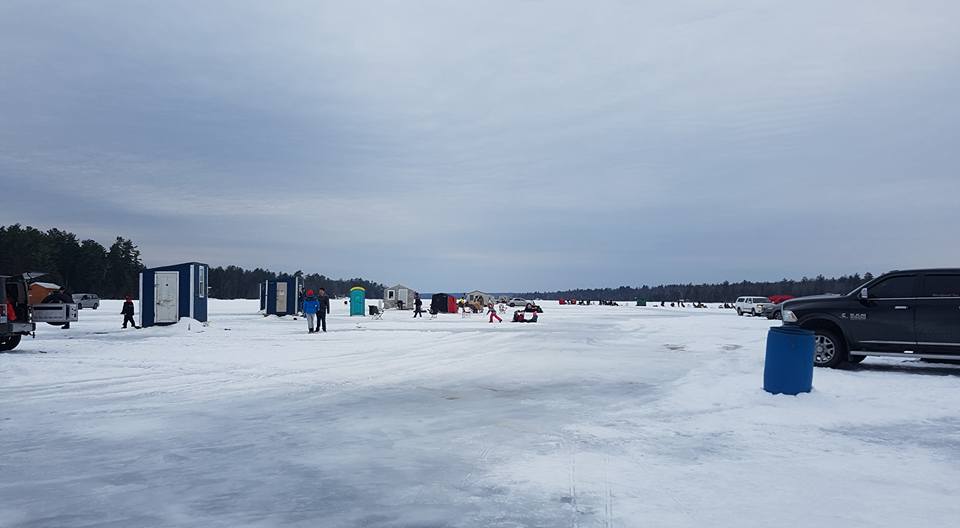 A wide expanse of frozen lake is dotted with ice shanties, vehicles, and groups of people enjoying a winter day outdoors. Portable restrooms and fishing shelters line the icy surface, while a cloudy sky stretches above.