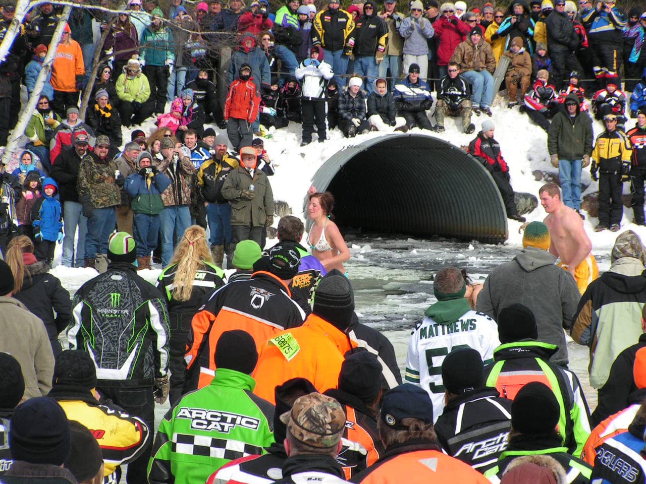 A large crowd gathers in winter gear to cheer on brave participants taking part in a polar plunge. Swimmers in swimsuits emerge from icy water flowing through a large culvert, while spectators line the snowy banks.