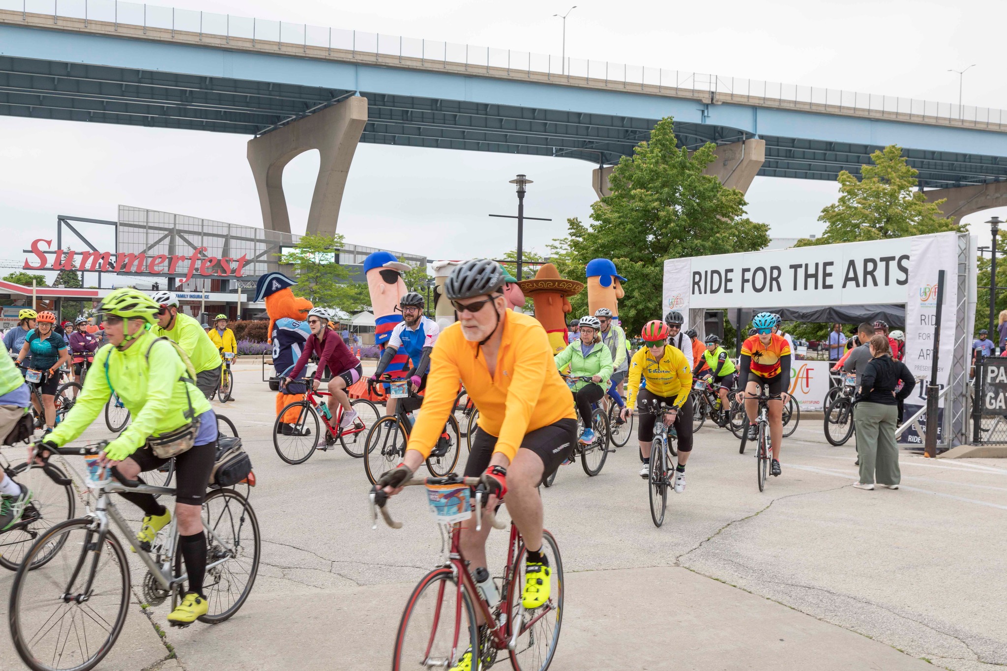 A large group of cyclists wearing bright jerseys ride together along a city street near an overpass during an organized biking event.