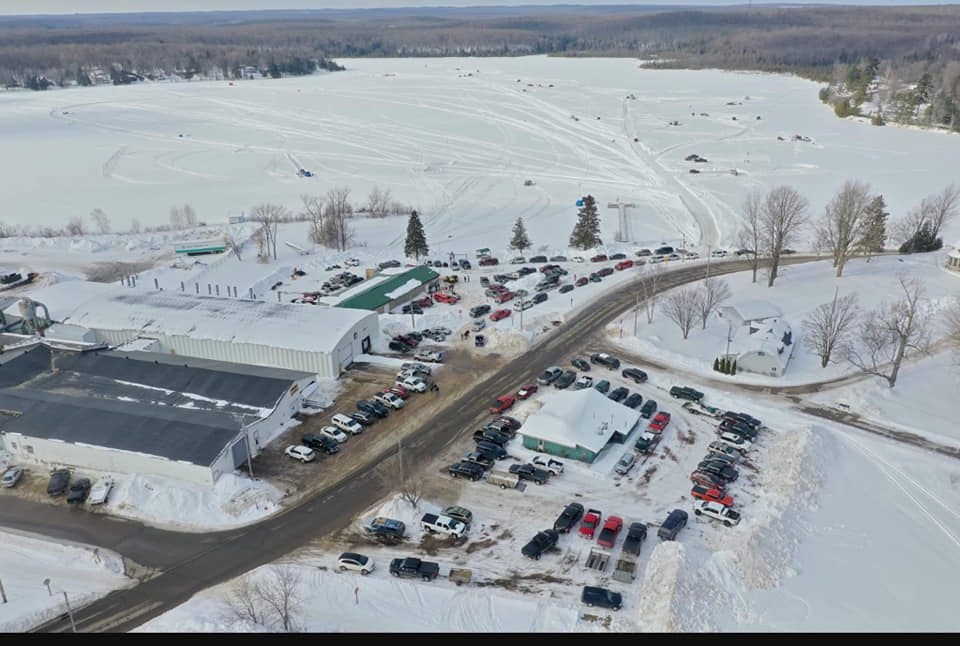 An aerial view of a bustling winter event shows a snow-covered lake filled with vehicles and tracks from snowmobiles or ATVs. Dozens of cars fill nearby parking lots and line the roads, while people gather around buildings and on the ice.