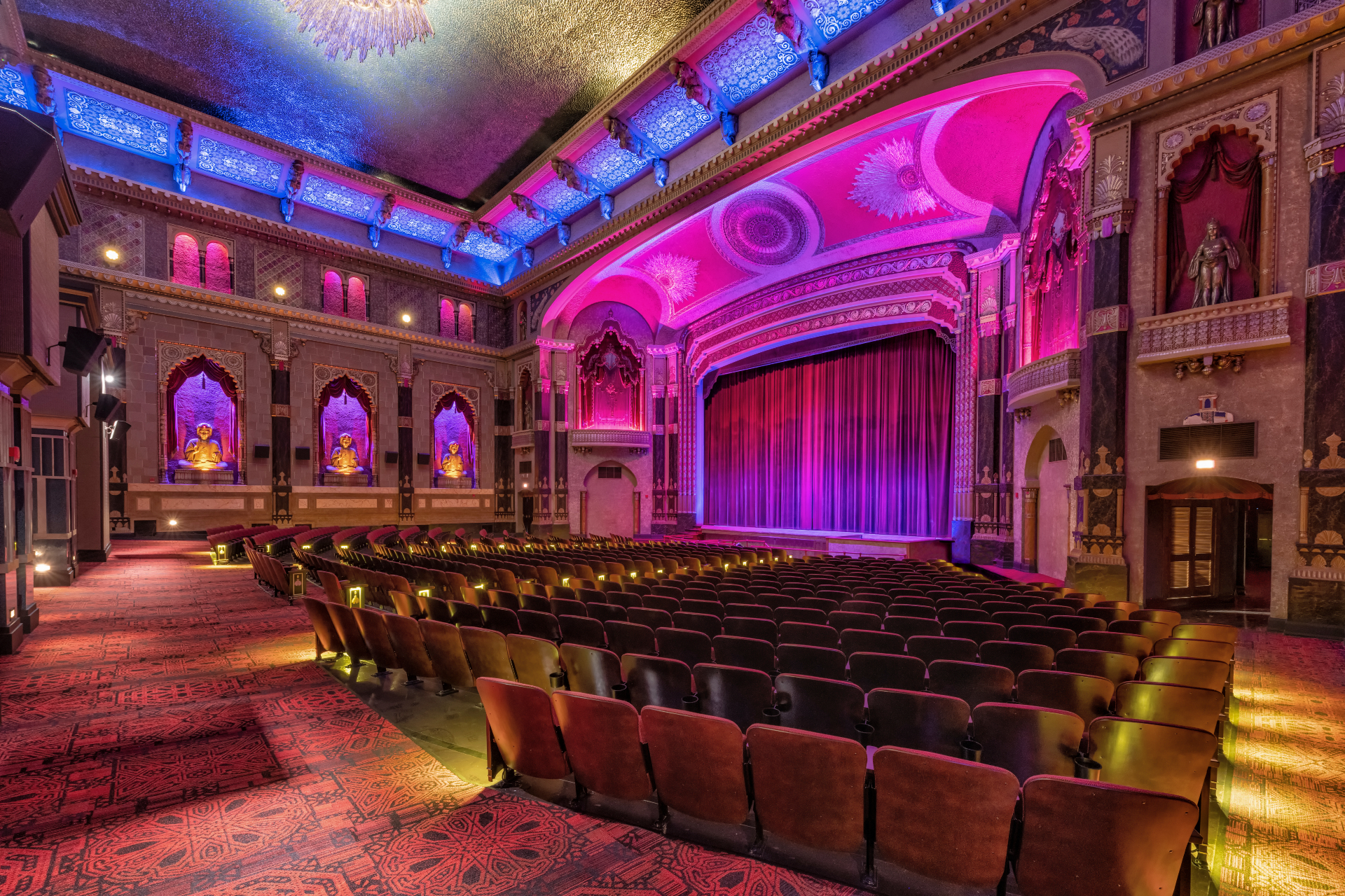 Interior of an ornate theater with colorful purple and blue lighting illuminating arched walls, decorative ceilings, and rows of red velvet seats.