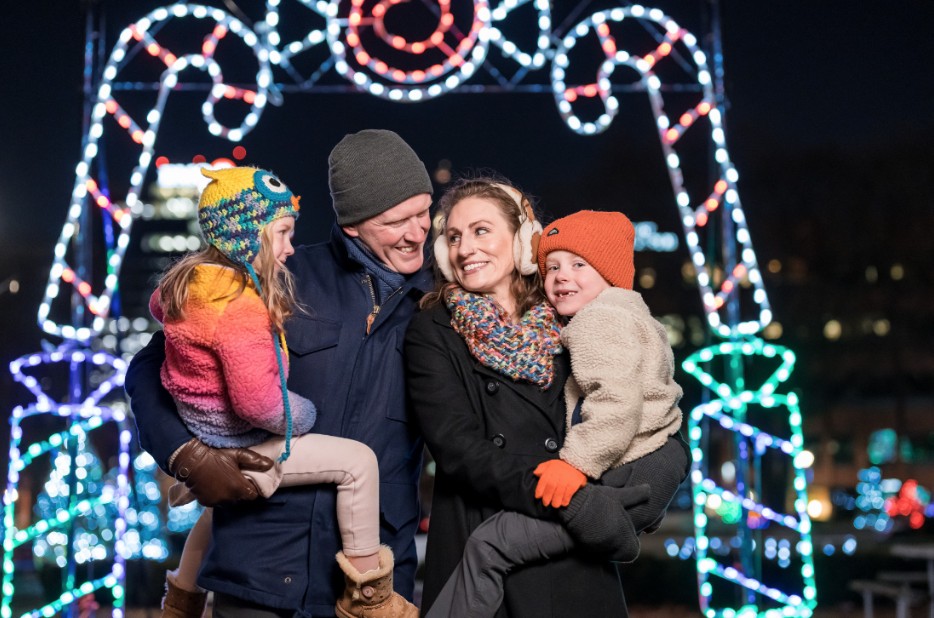 A bundled-up family smiles and poses together in front of a large illuminated candy cane light display during a holiday event at night.