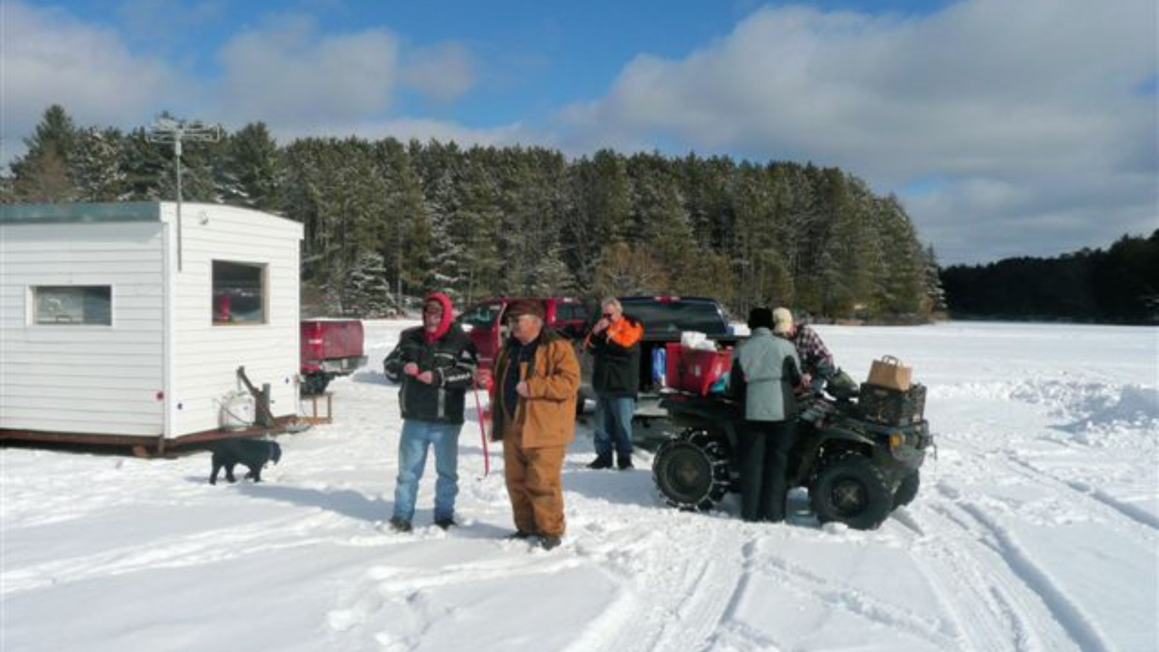 A group of people enjoys a sunny winter day on a frozen lake, gathered near an ice fishing shack, trucks, and an ATV loaded with supplies.