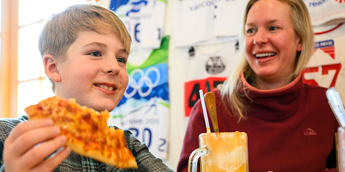Mother looking at son eating pizza with a milkshake drink on the table