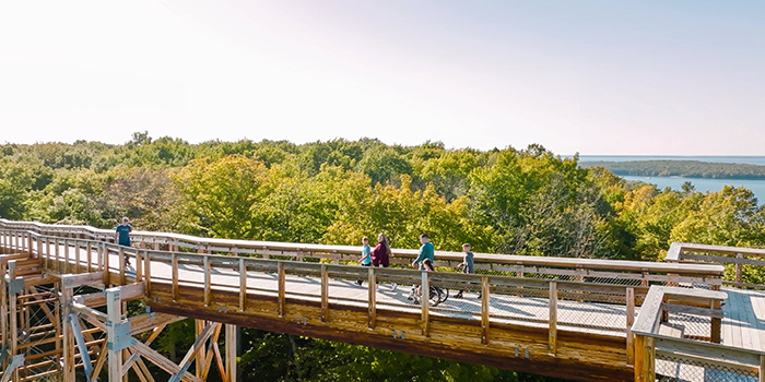 People walk down a wooden boardwalk backed by trees of fall colors and a lake