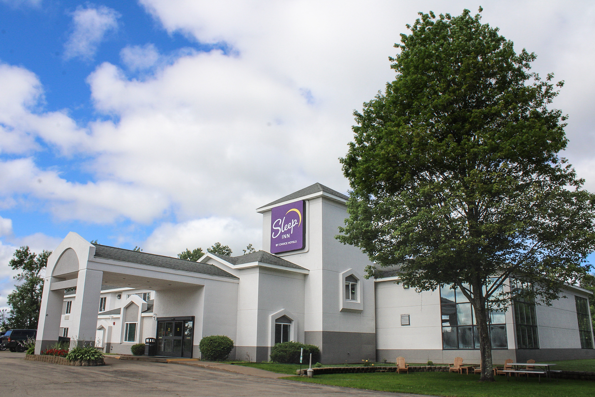 A two-story hotel building with a light-colored exterior and modern architectural design, featuring an arched entrance canopy and neatly landscaped flower beds. The parking lot is paved and bordered by trees in the background under a clear blue sky.