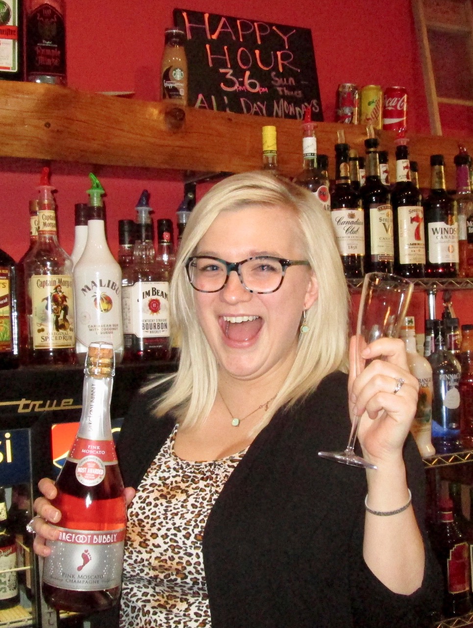 A cheerful bartender holding a bottle of sparkling pink Moscato and a champagne flute, standing in front of a fully stocked bar. A chalkboard sign behind her promotes happy hour specials, adding a fun and festive vibe.
