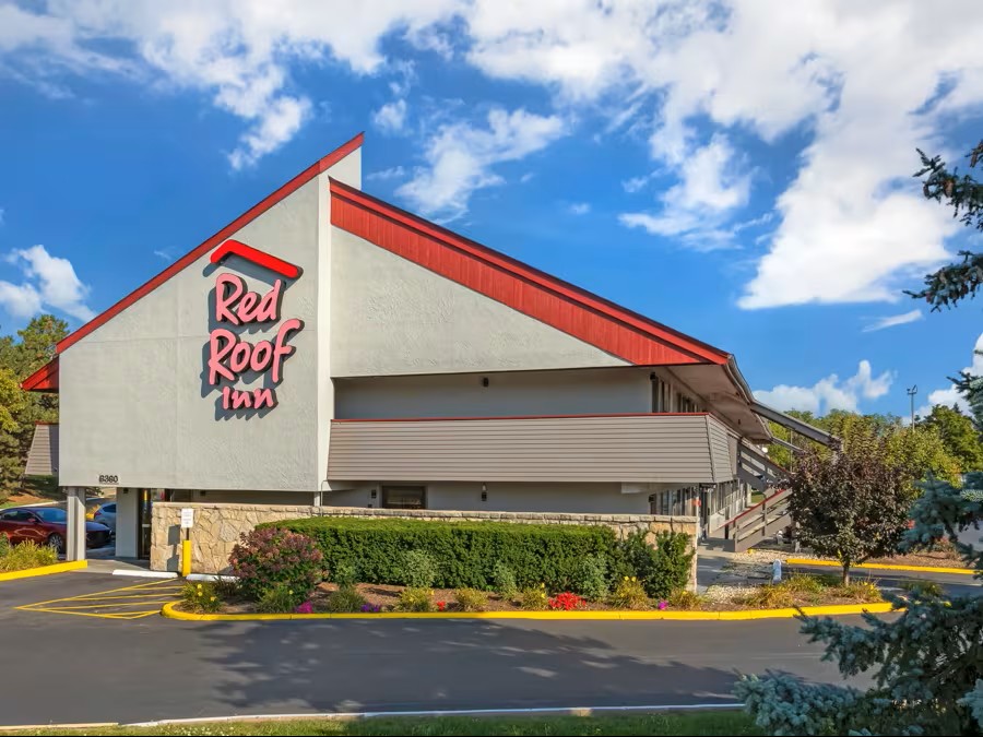 Exterior of a Red Roof Inn hotel with a sloped roof and large logo on the side, surrounded by a parking lot and landscaping under a blue sky with clouds.