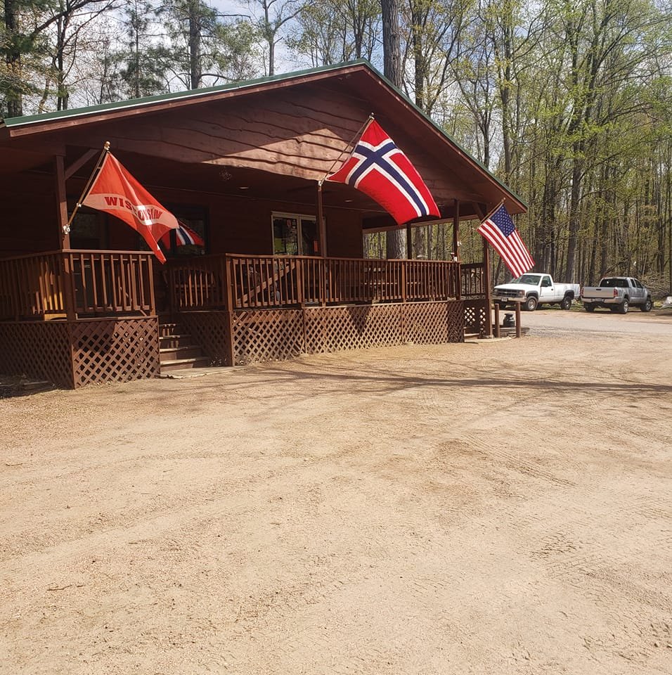 A rustic log cabin-style building with a wraparound porch and three flags—Wisconsin, Norway, and the United States—hanging from the roofline. The building sits among tall trees on a dirt lot with a few pickup trucks parked nearby, creating a warm and welcoming woodland setting.