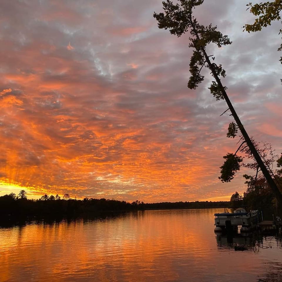 A breathtaking view of a lake at sunset, with vivid orange and pink hues reflecting off the water beneath a dramatic, cloud-filled sky. A pontoon boat is docked at the edge of the frame, and tall trees are silhouetted along the shoreline, adding to the serene and picturesque scene.