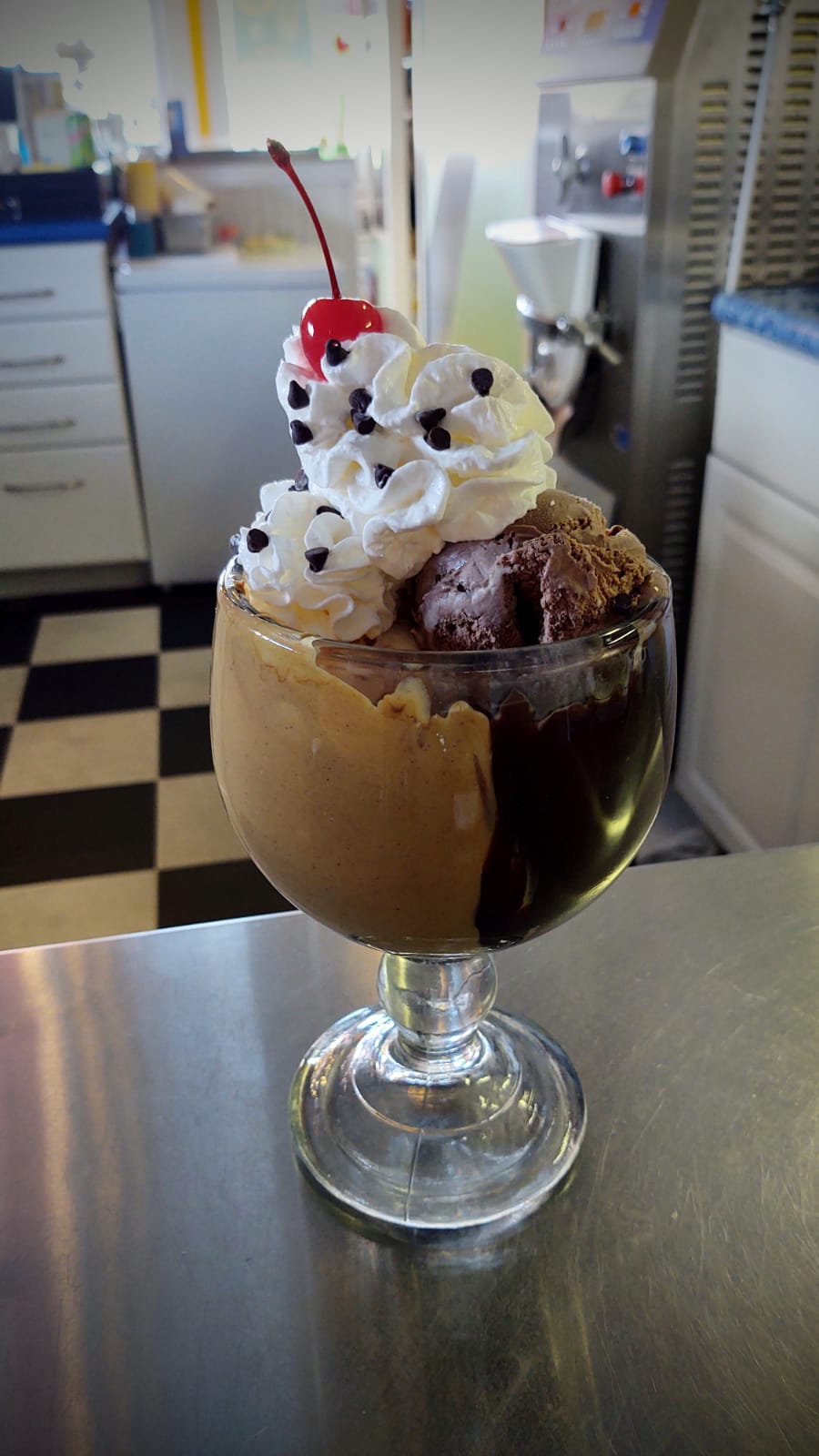 A rich, chocolate ice cream sundae is served in a glass goblet, layered with peanut butter sauce, whipped cream, mini chocolate chips, and topped with a cherry.
