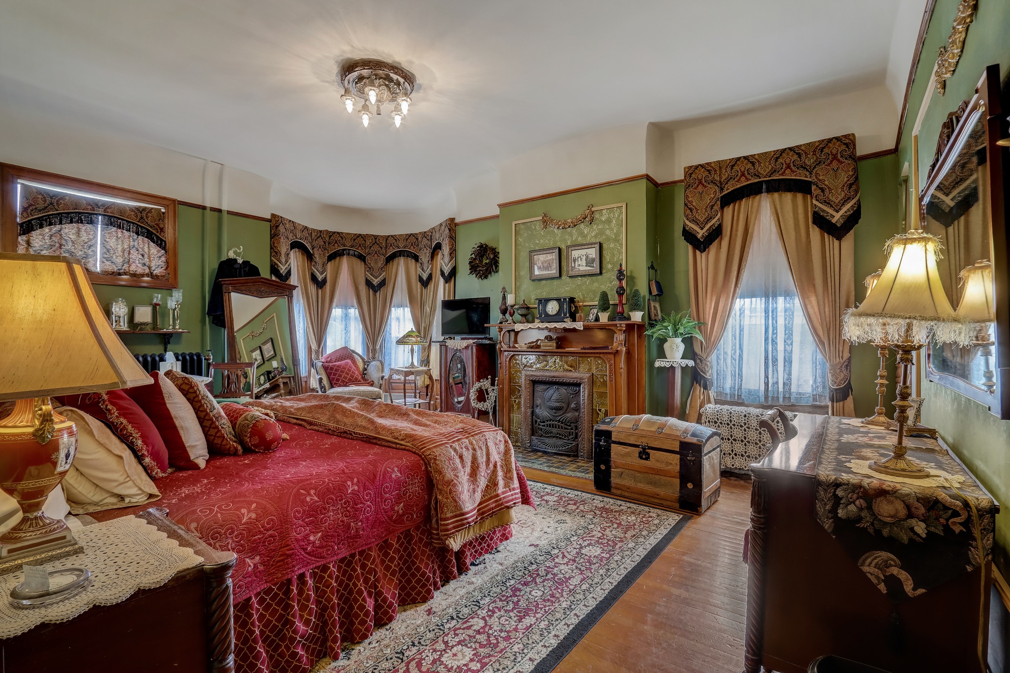 A cozy Victorian-style bedroom with rich red bedding, antique furniture, floral decor, and a chandelier, creating a warm and elegant atmosphere.