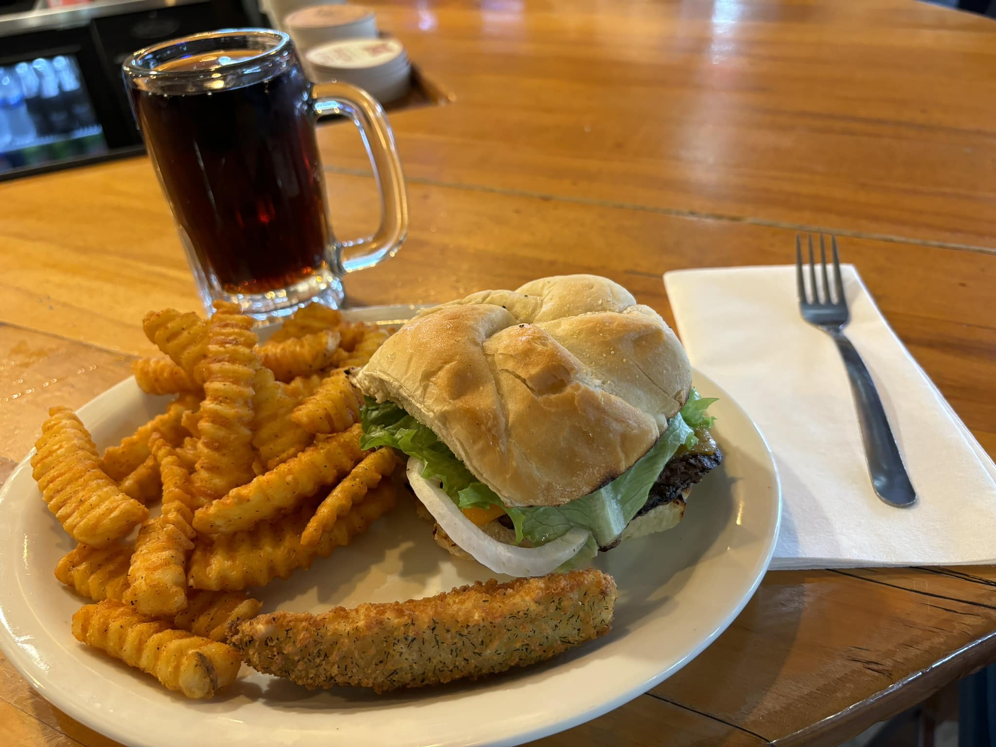 A classic cheeseburger with lettuce and onion is paired with crispy crinkle-cut fries, a fried pickle spear, and an ice-cold root beer—perfect comfort food at the bar inside Foxwood Tavern.