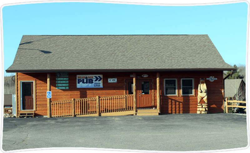 A wooden log-style building with a covered front porch and handicap-accessible ramp. Signs on the front identify it as a pub with food and spirits, and a carved bear statue stands near the entrance. The building sits against a clear sky backdrop with ample parking.
