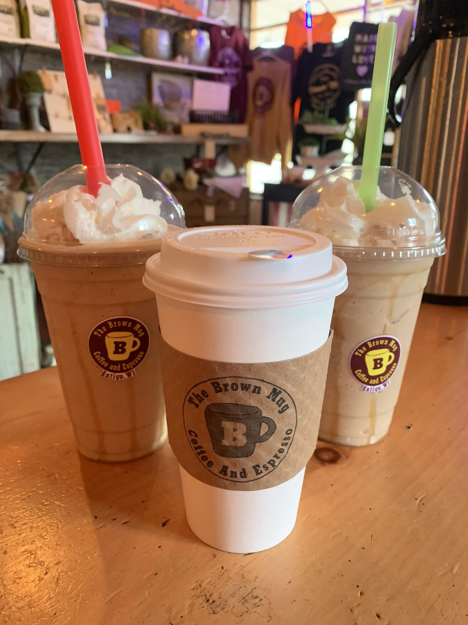 Three handcrafted coffee drinks from The Brown Mug Coffee and Espresso in Antigo, WI sit on a cozy wooden counter—two iced drinks topped with whipped cream and one steaming hot beverage, ready to warm or energize any visitor exploring Langlade County.