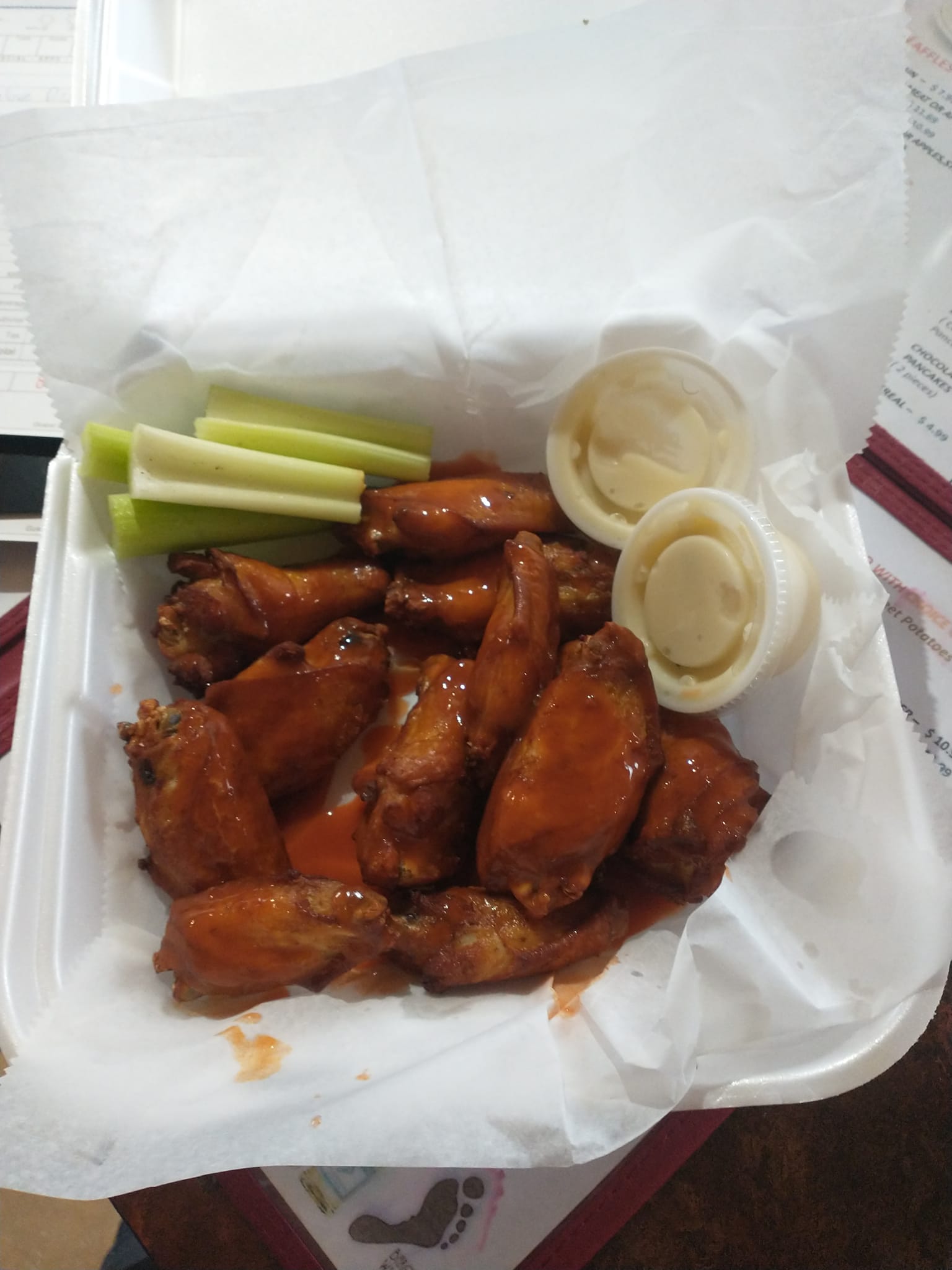 A takeout container filled with saucy buffalo chicken wings, served with celery sticks and two cups of ranch or blue cheese dressing.
