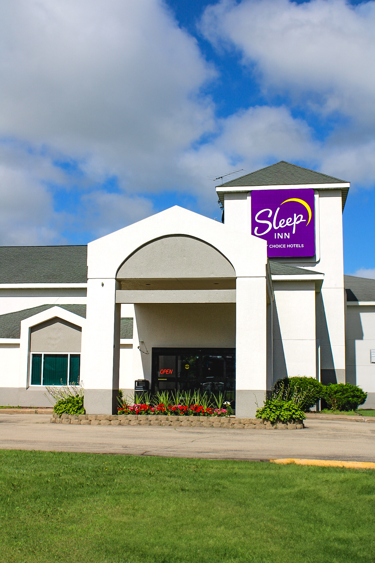 A bright and welcoming exterior shot of the Sleep Inn hotel in Antigo, WI, under a partly cloudy blue sky. The clean, white building features a modern design with a large entry canopy and the signature purple Sleep Inn sign above. Fresh landscaping with flowers and greenery surrounds the entrance, inviting travelers to enjoy a comfortable and convenient stay.