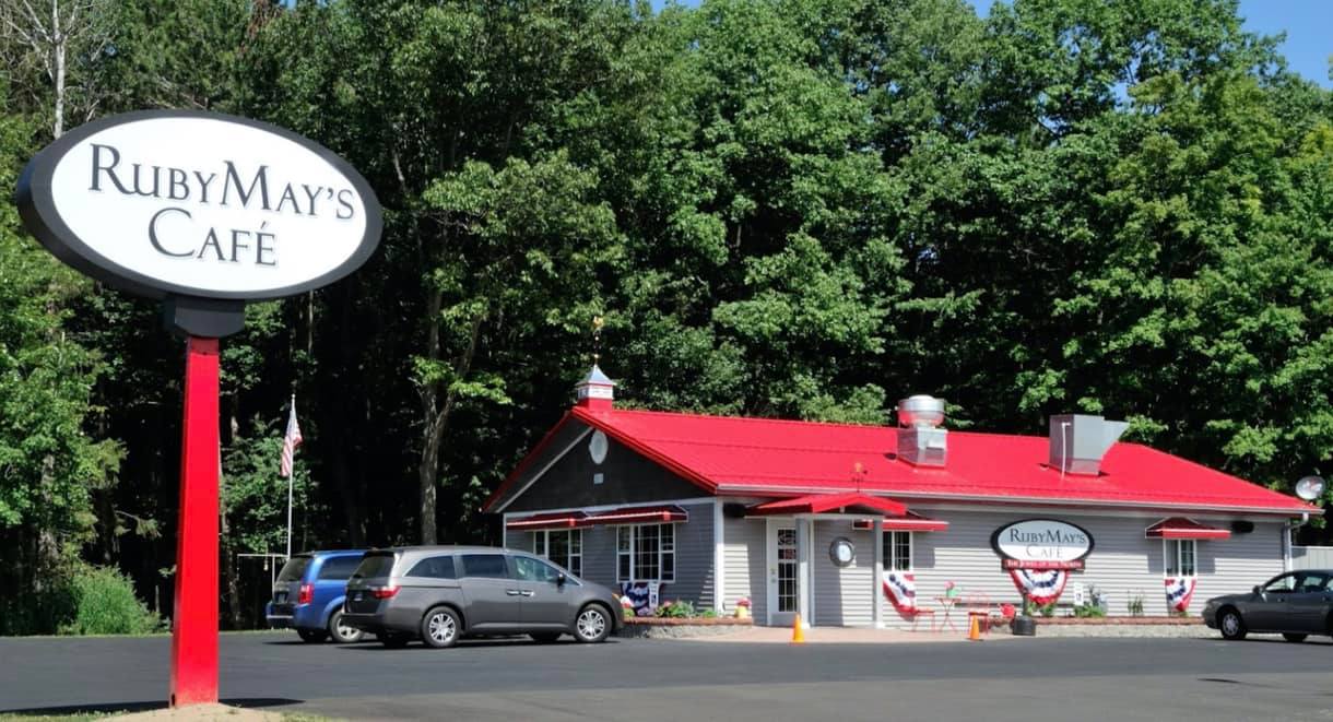 A cozy café building with a bright red roof and gray siding, nestled against a wooded backdrop. The parking lot is partially filled with vehicles, and a large oval sign reading “Ruby May’s Café” stands at the entrance.