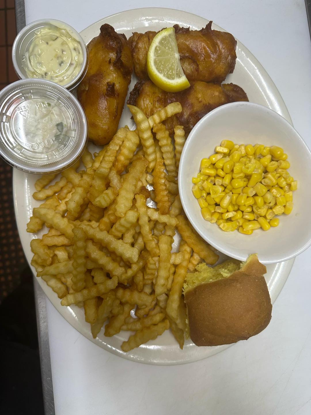 A hearty meal featuring golden crinkle-cut fries, breaded fish fillets topped with a lemon wedge, a dinner roll, corn, and two containers of tartar sauce.