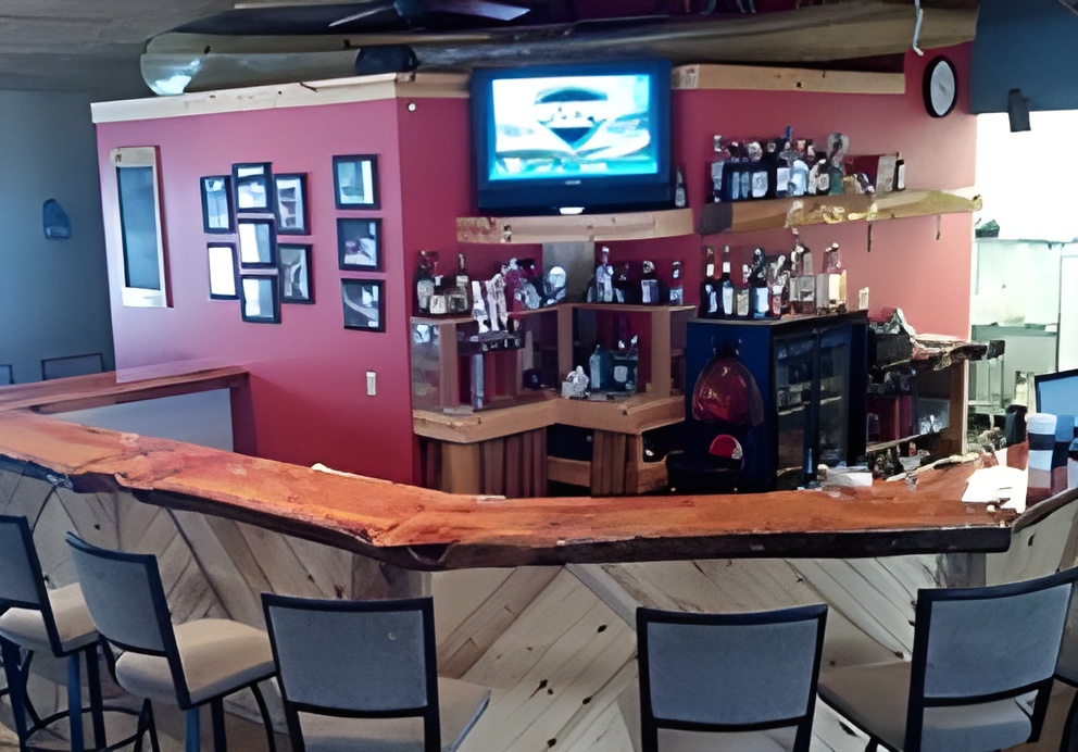 A cozy bar area with a live-edge wood countertop and high-backed chairs, set against a deep red wall. Shelves are lined with liquor bottles, and a TV is mounted above. The space combines rustic charm with a casual atmosphere.