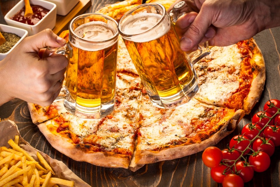 Two mugs of beer clink over a freshly baked pizza—celebrating good times, good food, and the laid-back charm of dining at Foxwood Tavern.