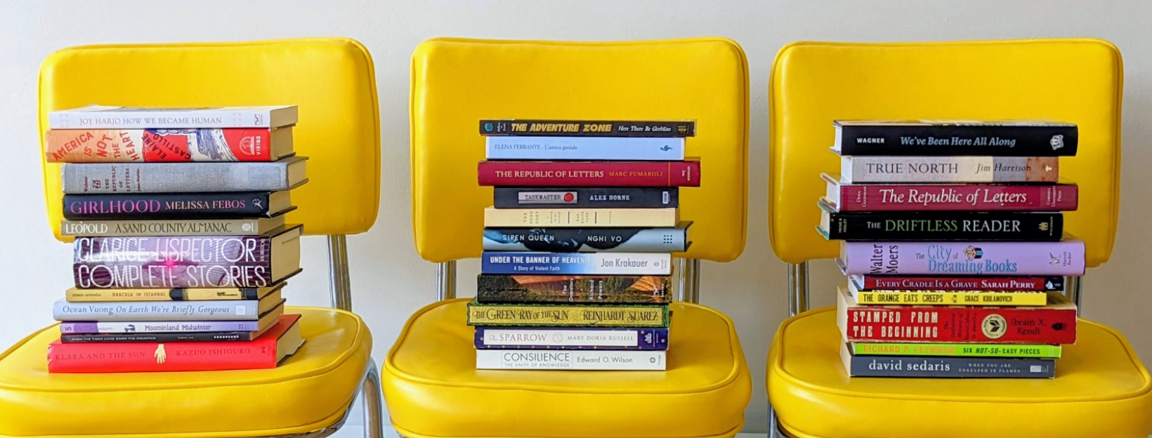 A row of three yellow chairs, each with a stack of books in various sizes and colors.