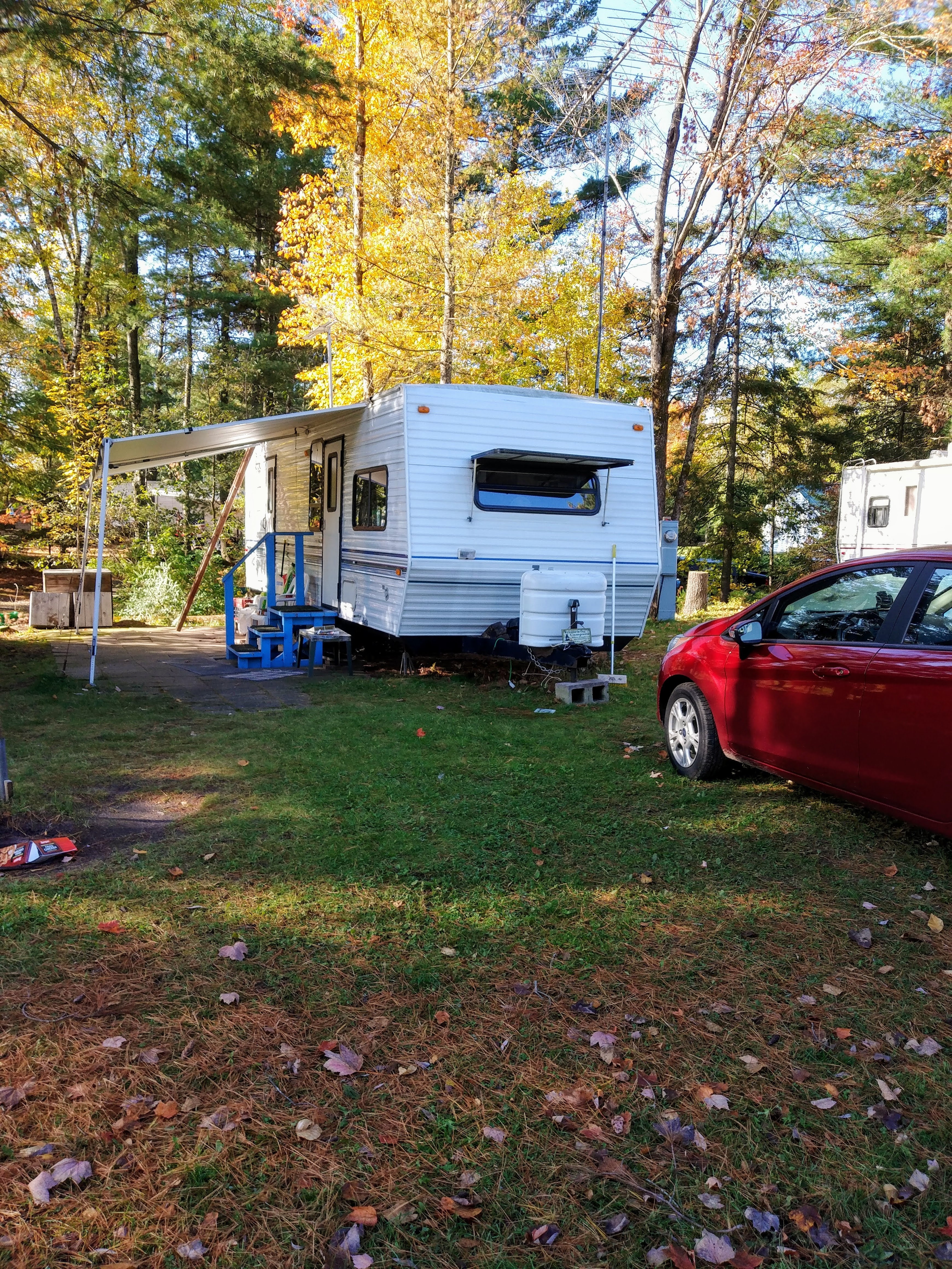 A white travel trailer with an awning is parked on a grassy campsite surrounded by tall autumn-colored trees. A small blue staircase leads to the trailer door, and a red car is parked nearby. The area is shaded and serene, with signs of fall leaves scattered across the ground.