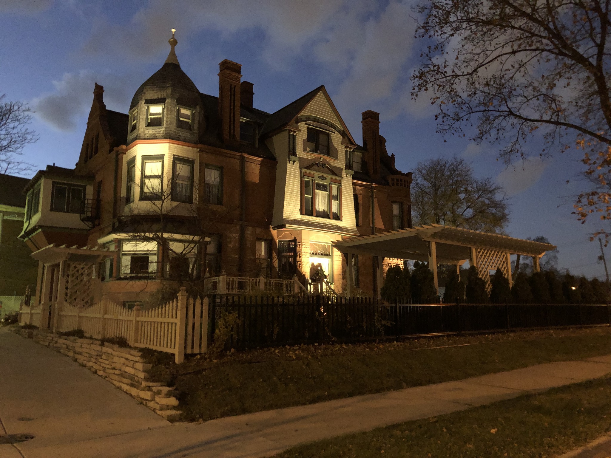A large historic Victorian-style house illuminated with warm lights at dusk, showcasing ornate architecture and a wraparound porch.