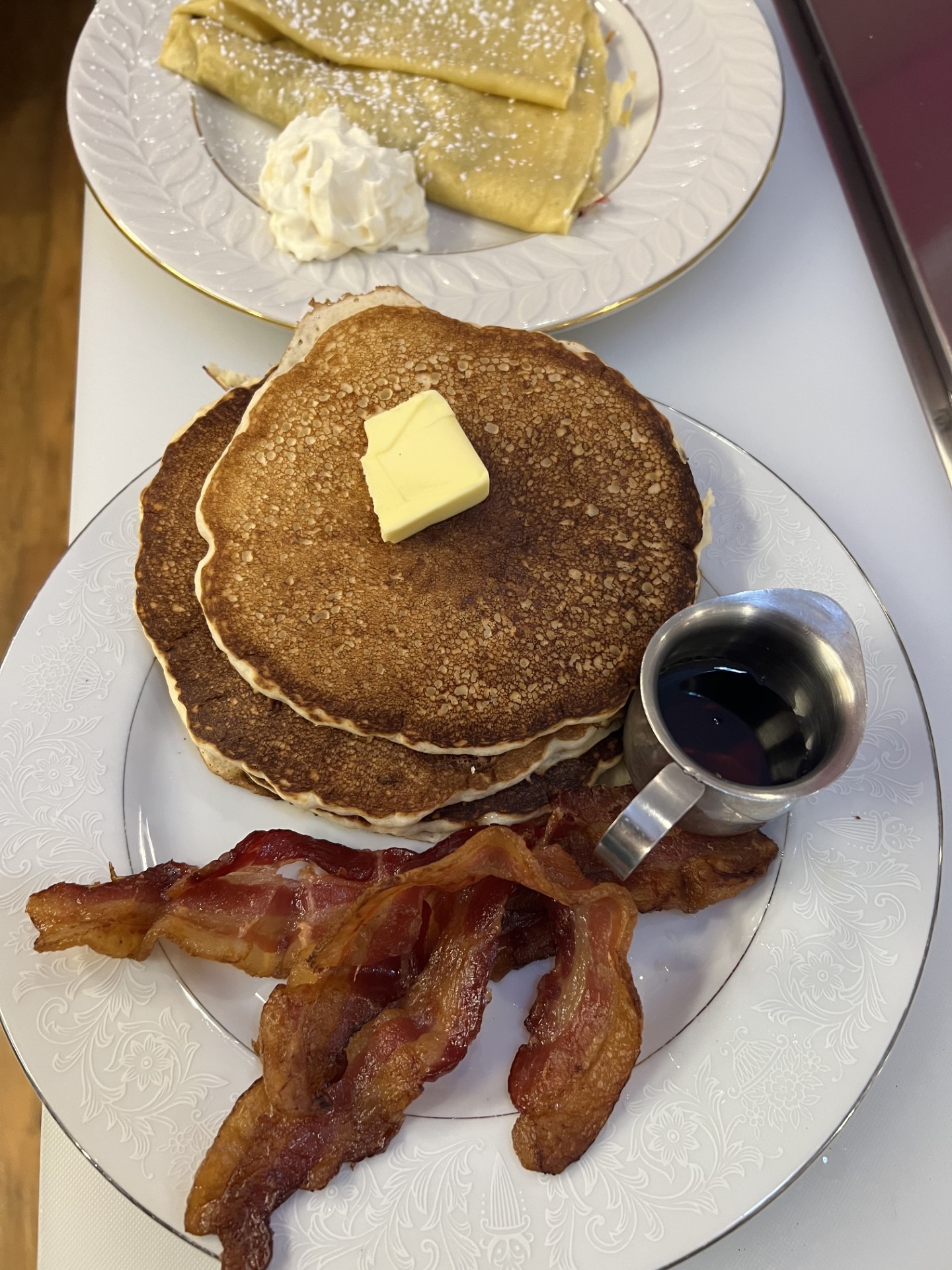 crepes, pancakes, and bacon on a plate with butter, whipped cream, and maple syrup.